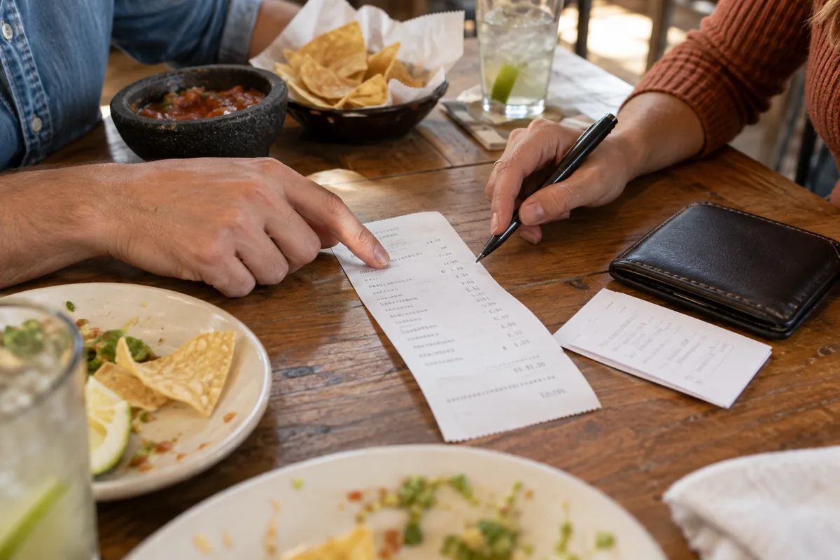 Two diners review a generic itemized restaurant receipt and payment slip beside tortilla chips, salsa and a closed wallet